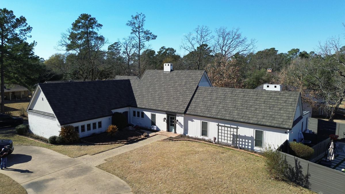 Drone view of a long single-story white house with a dark roof, set among tall pines under a clear sky