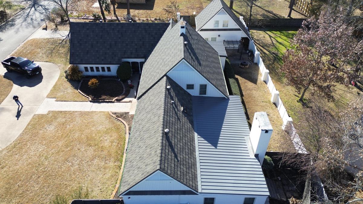 Overhead drone view of a white house with a dark roof and a light gray paneled roof section along one wing