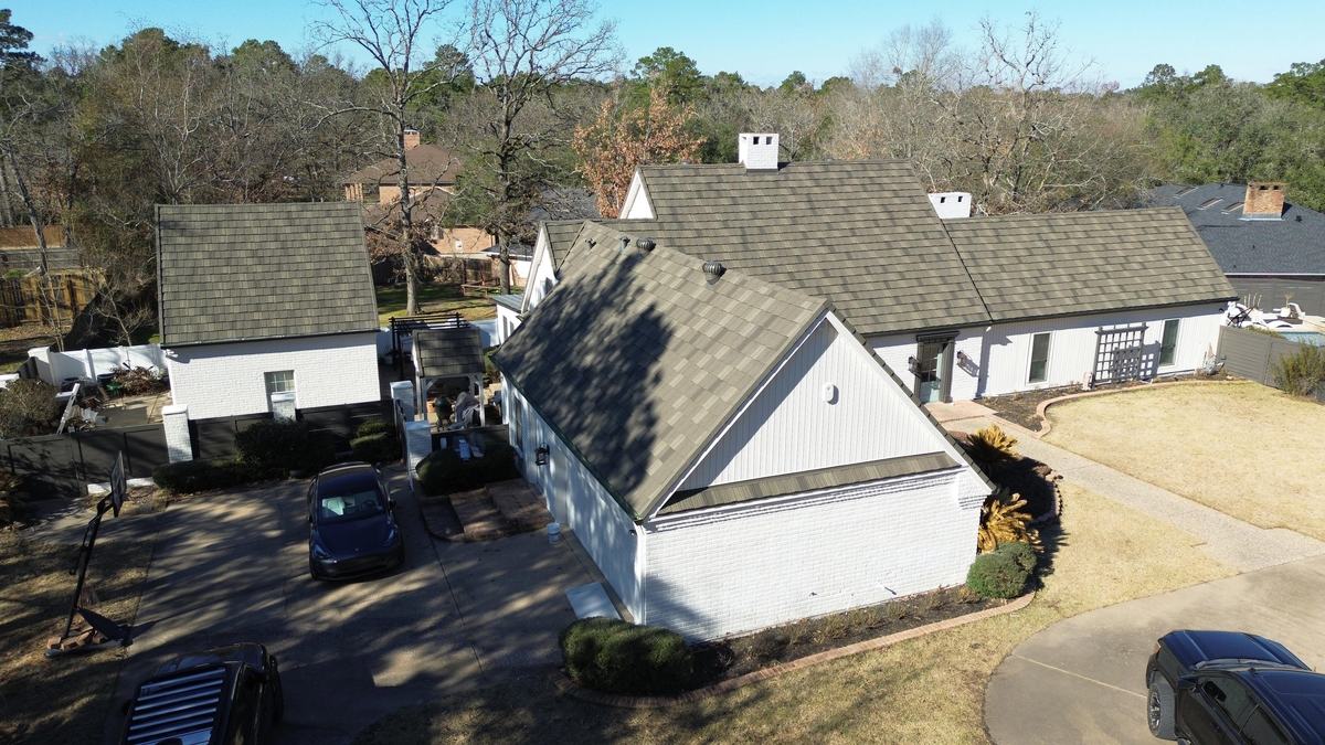 Drone view of a white house with a gray-brown roof, surrounded by bare winter trees