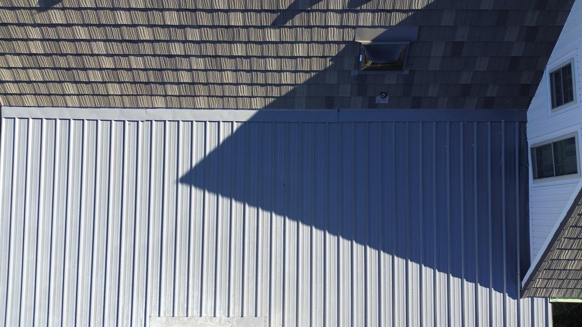 Close drone view where a light gray paneled roof section meets a textured dark roof