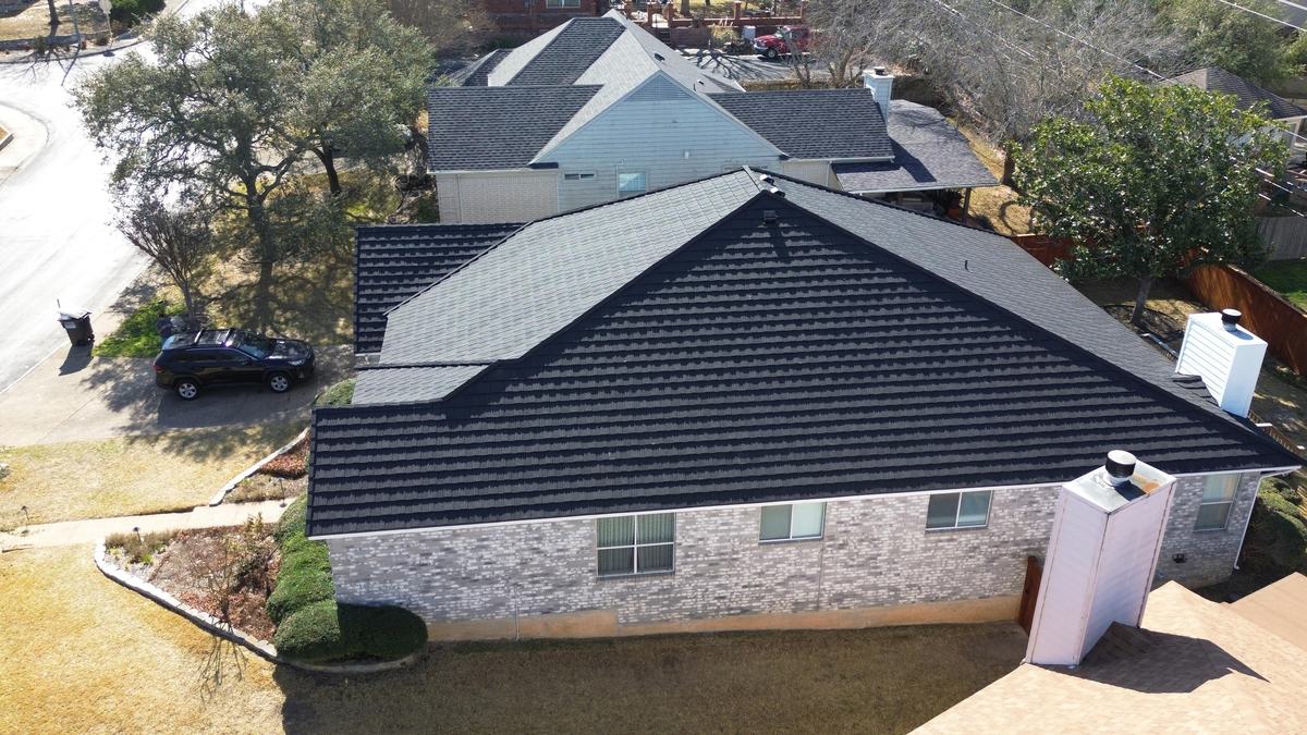 Rear drone view of a brick home with a charcoal roof and white chimney near a tree-lined street