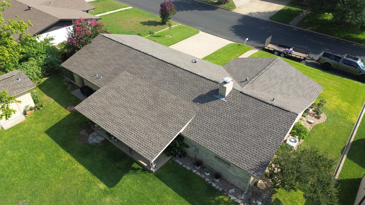 Angled overhead drone view of a ranch home with a brown-gray roof and a green lawn