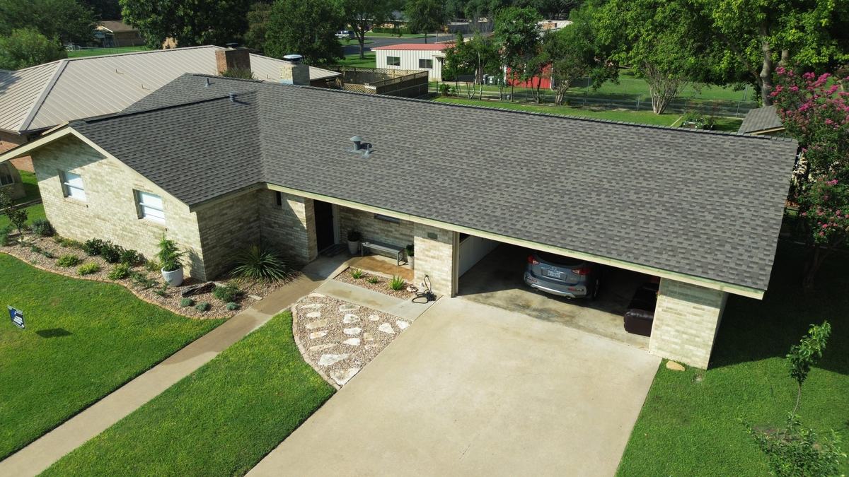 Drone view of a stone ranch home with a gray roof and a covered carport